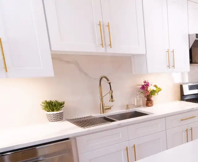 White shaker cabinets with gold pulls, a white countertop and a matching marble-look slab backsplash behind a gold faucet